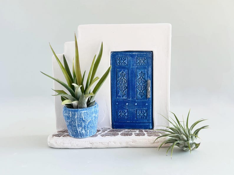 Pode incluir: Uma estrutura de cer&acirc;mica branca com uma porta azul apresentando padr&otilde;es em relevo e um puxador prateado. Um vaso azul texturizado contendo uma suculenta verde repousa na base, acompanhado por uma pequena planta a&eacute;rea no ch&atilde;o.