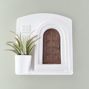 May include: White wall-mounted planter with a miniature arched doorway and a small potted succulent. The door is brown with a dark handle. The planter has a textured surface and a small shelf.