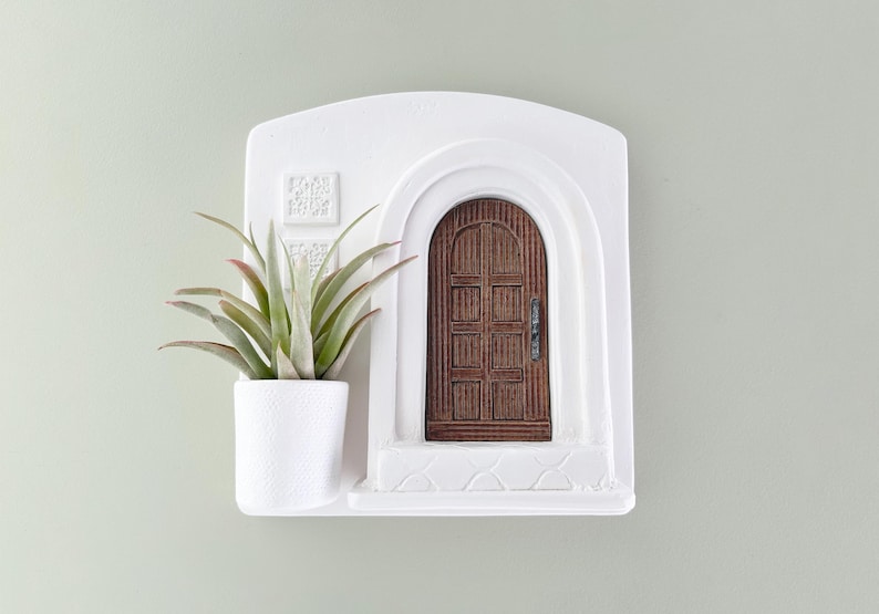 May include: White wall-mounted planter with a miniature arched doorway and a small potted succulent. The door is brown with a dark handle. The planter has a textured surface and a small shelf.
