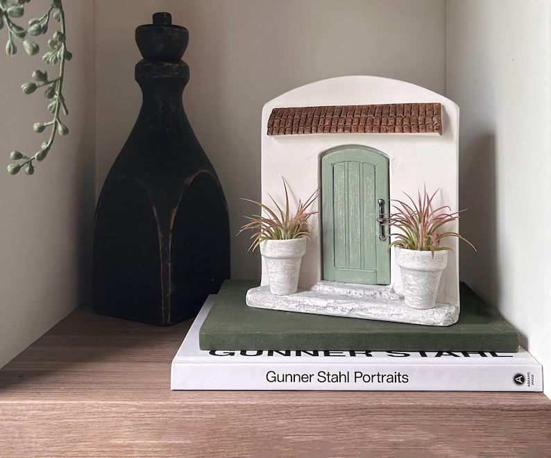 Pu&ograve; includere: Una porticina decorativa con una porta verde, un telaio bianco e un tetto di tegole marroni. Due piccole piante in vaso affiancano la porta. La porta &egrave; posizionata su un blocco verde sopra un libro intitolato "Gunner Stahl Portraits". A sinistra c'&egrave; una bottiglia decorativa alta e nera.