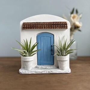 Pu&ograve; includere: Una facciata in stucco bianco in miniatura con una porta blu e un tetto di tegole marroni. Due piccole piante in vaso fiancheggiano la porta. La scena evoca un'estetica mediterranea o toscana, ideale per l'arredamento della casa.