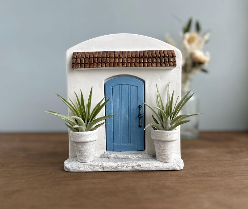 Pu&ograve; includere: Una facciata in stucco bianco in miniatura con una porta blu e un tetto di tegole marroni. Due piccole piante in vaso fiancheggiano la porta. La scena evoca un'estetica mediterranea o toscana, ideale per l'arredamento della casa.