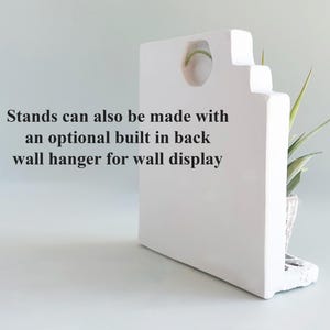 Pode incluir: Um suporte retangular branco com um recorte circular no topo e um pequeno suporte para plantas na lateral. O texto no suporte diz: Stands can also be made with an optional built in back wall hanger for wall display.