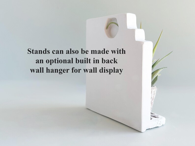 Pode incluir: Um suporte retangular branco com um recorte circular no topo e um pequeno suporte para plantas na lateral. O texto no suporte diz: Stands can also be made with an optional built in back wall hanger for wall display.