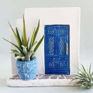 Pode incluir: Uma escultura arquitet&oacute;nica branca com uma porta azul com entalhes ornamentados e um puxador prateado. Um vaso azul texturizado com uma suculenta verde assenta numa base de pedra ao lado de uma pequena planta a&eacute;rea &agrave; direita.