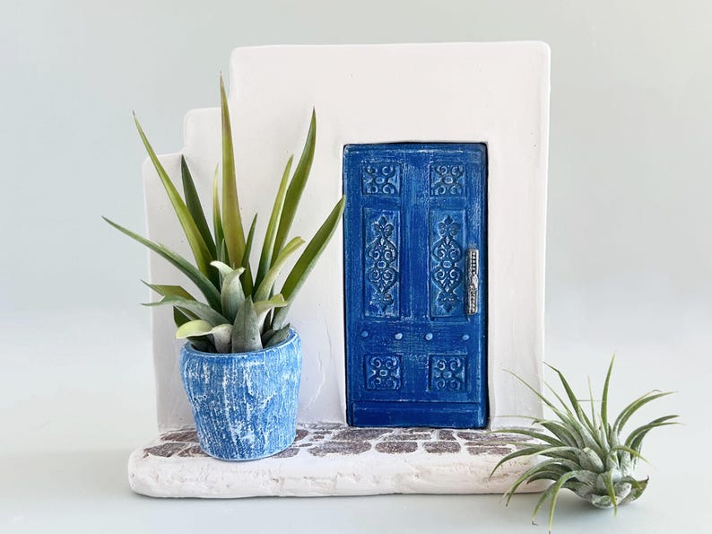Pode incluir: Uma escultura arquitet&oacute;nica branca com uma porta azul com entalhes ornamentados e um puxador prateado. Um vaso azul texturizado com uma suculenta verde assenta numa base de pedra ao lado de uma pequena planta a&eacute;rea &agrave; direita.