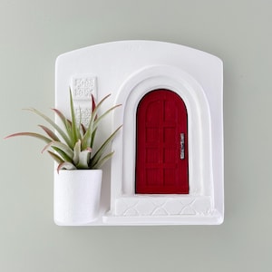 May include: A white wall-mounted planter with a red door design. The planter includes a small white pot holding a green and red air plant. The door has a silver handle and is set in an arched doorway.