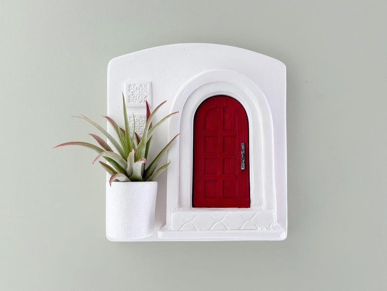 May include: A white wall-mounted planter with a red door design. The planter includes a small white pot holding a green and red air plant. The door has a silver handle and is set in an arched doorway.