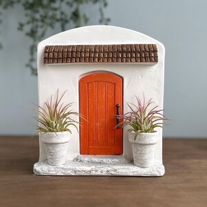 Pu&ograve; includere: Una facciata in stucco bianco in miniatura con una porta arancione, un tetto di tegole marroni e due piante in vaso. La porta ha una maniglia scura. Le piante hanno foglie verdi e rosa. La facciata &egrave; su una superficie di legno.