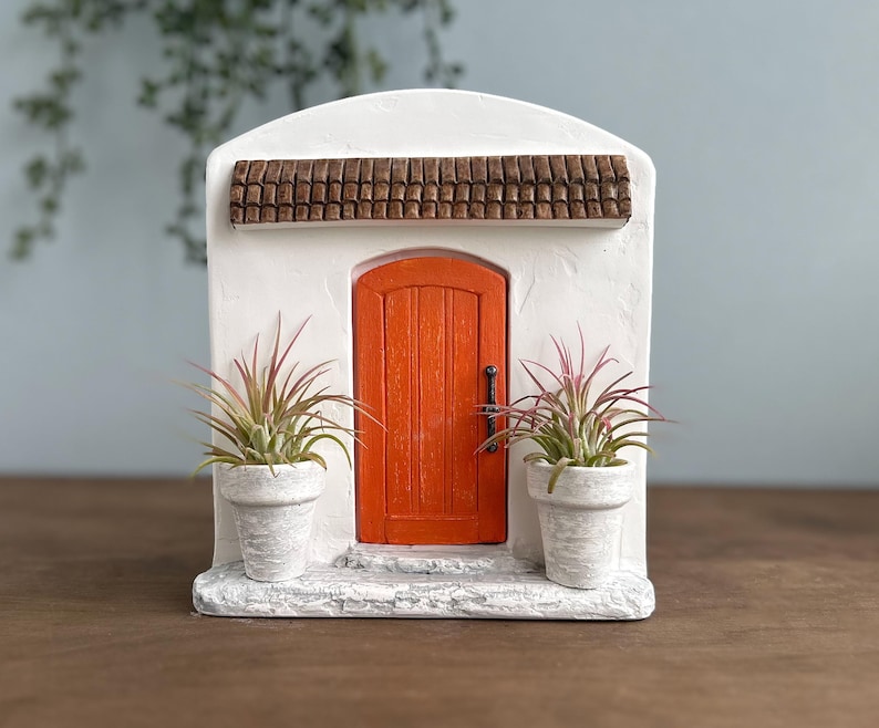Pu&ograve; includere: Una facciata in stucco bianco in miniatura con una porta arancione, un tetto di tegole marroni e due piante in vaso. La porta ha una maniglia scura. Le piante hanno foglie verdi e rosa. La facciata &egrave; su una superficie di legno.