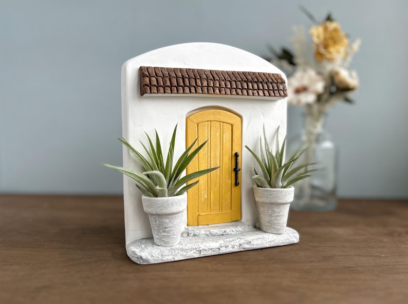 Pu&ograve; includere: Una facciata in stucco bianco in miniatura con una porta gialla, un tetto in tegole marroni e due piante in vaso. La porta ha una maniglia nera. Le piante hanno lunghe foglie verdi. Un oggetto decorativo.