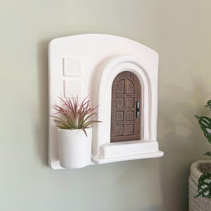 May include: A white wall-mounted decorative piece featuring a miniature arched doorway with a brown wooden door. A small white pot with a plant with pink and green leaves sits below the door. Two square decorative elements are above the plant.