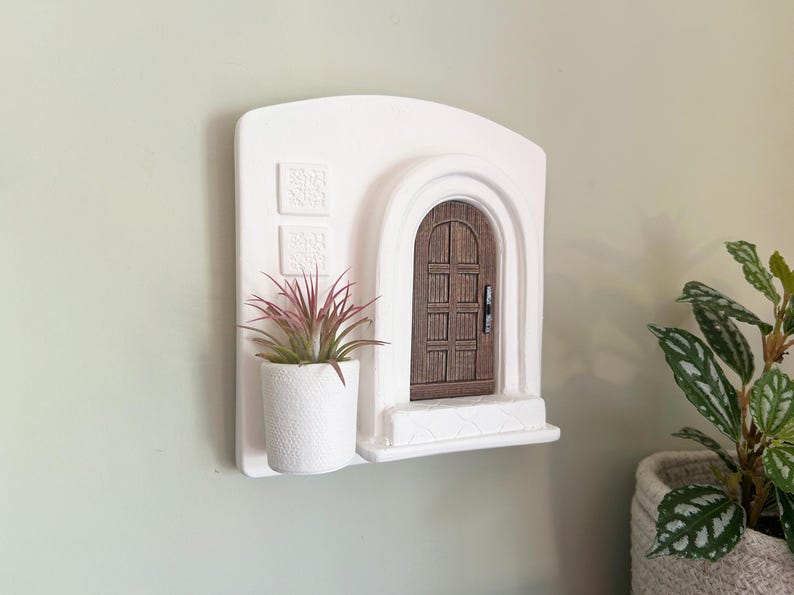 May include: A white wall-mounted decorative piece featuring a miniature arched doorway with a brown wooden door. A small white pot with a plant with pink and green leaves sits below the door. Two square decorative elements are above the plant.