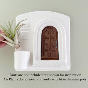 May include: White wall-mounted planter with a miniature arched doorway and a small white pot. A brown wooden door is set within the arch. The planter is designed to hold air plants, which are shown for inspiration. The text says, "Plants are not included but shown for inspiration. Air Plants do not need soil and easily fit in the mini pots."