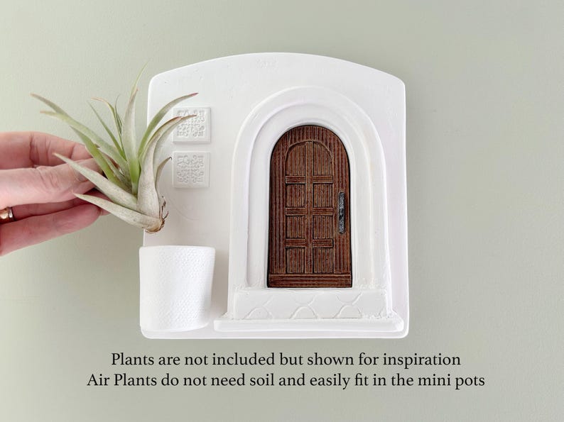 May include: White wall-mounted planter with a miniature arched doorway and a small white pot. A brown wooden door is set within the arch. The planter is designed to hold air plants, which are shown for inspiration. The text says, "Plants are not included but shown for inspiration. Air Plants do not need soil and easily fit in the mini pots."