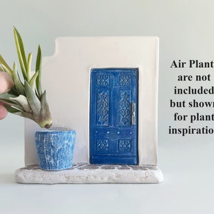 Pode incluir: Uma escultura de parede branca com uma porta azul com entalhes intrincados e um vaso azul texturizado a condizer. O texto &agrave; direita diz: Air Plants are not included but shown for plant inspiration. A base tem uma textura de pedra.