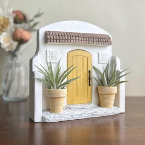 May include: A miniature white stucco facade with a yellow door, brown tile roof, and two potted plants. The decorative piece has a stone step and is designed to resemble a small house or building.