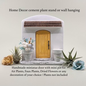 Pu&ograve; includere: Una porta in miniatura in cemento con una porta gialla e un tetto marrone, progettata per l'arredamento della casa. Include un piccolo vaso per piante aeree, piante finte o fiori secchi. Il testo recita: "Home Decor cement plant stand or wall hanging" e "Handmade miniature door with mini pot for Air Plants, Faux Plants, Dried Flowers or any decoration of your choice / Plants not included."