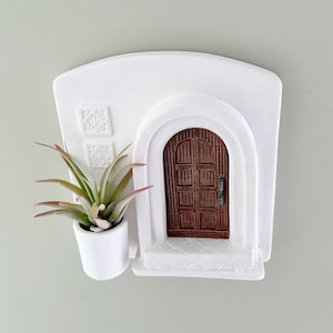 May include: A white wall-mounted planter shaped like a Mediterranean-style building with a brown wooden door. A small air plant with green and red-tipped leaves sits in a white pot attached to the side. Decorative squares are above the door.