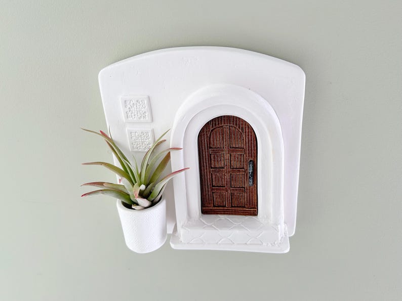 May include: A white wall-mounted planter shaped like a Mediterranean-style building with a brown wooden door. A small air plant with green and red-tipped leaves sits in a white pot attached to the side. Decorative squares are above the door.