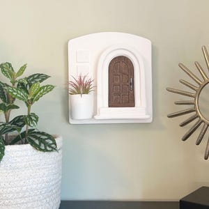 May include: A white wall-mounted decorative piece featuring a miniature arched brown door. A small white planter with a red and green plant sits to the left of the door. A gold sunburst sculpture and a potted plant are also visible.
