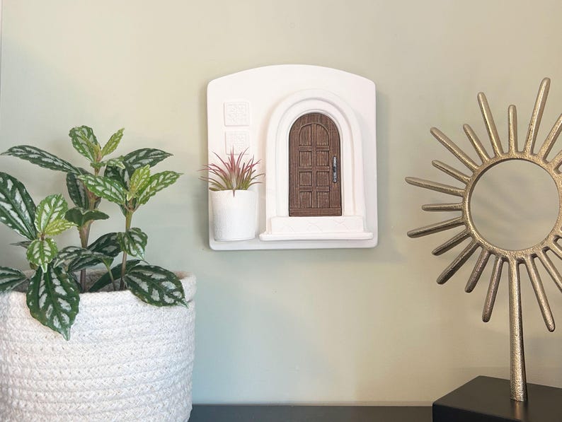 May include: A white wall-mounted decorative piece featuring a miniature arched brown door. A small white planter with a red and green plant sits to the left of the door. A gold sunburst sculpture and a potted plant are also visible.