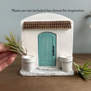 Pu&ograve; includere: Un edificio in miniatura in stucco bianco con una porta turchese, un tetto in tegole marroni e due piccoli vasi bianchi. Il testo "Plants are not included but shown for inspiration" &egrave; sopra l'edificio. Una mano tiene una pianta vicino al vaso.
