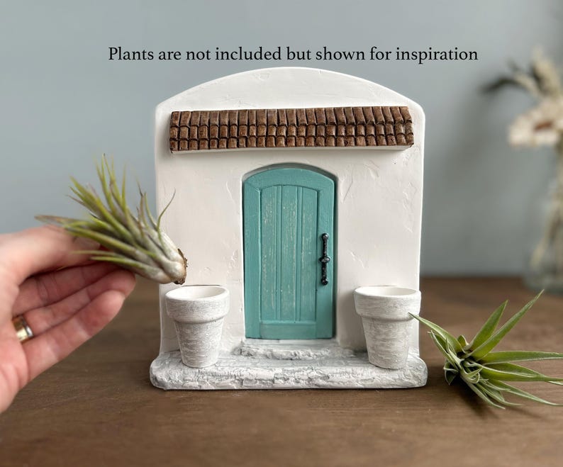 Pu&ograve; includere: Un edificio in miniatura in stucco bianco con una porta turchese, un tetto in tegole marroni e due piccoli vasi bianchi. Il testo "Plants are not included but shown for inspiration" &egrave; sopra l'edificio. Una mano tiene una pianta vicino al vaso.