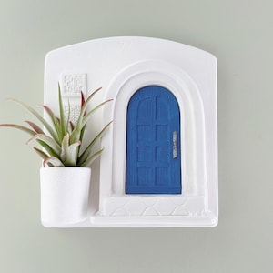 May include: A white wall-mounted planter with a blue door design and a small white pot holding a green and red-tipped plant. The planter has a textured surface and is mounted on a light green wall.