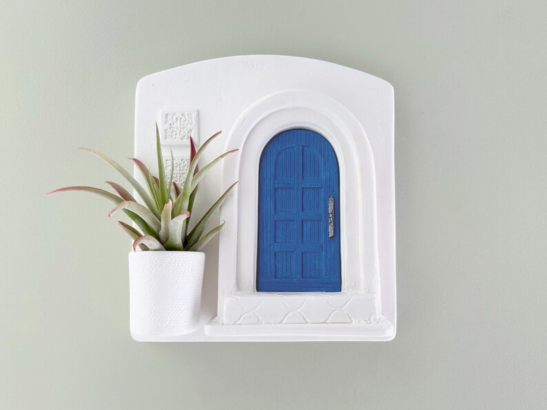 May include: A white wall-mounted planter with a blue door design and a small white pot holding a green and red-tipped plant. The planter has a textured surface and is mounted on a light green wall.