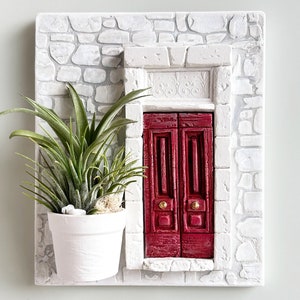 May include: A white ceramic wall decor piece featuring a red double door with gold accents, set in a white stone wall with a green plant in a white pot.