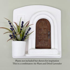 May include: A white wall decor piece featuring a miniature arched doorway with a brown wooden door. A white planter with a green plant and purple flowers sits to the left. The text at the bottom says, "Plants not included but shown for inspiration. This is a combinaton Air Plant and Dried Lavender."