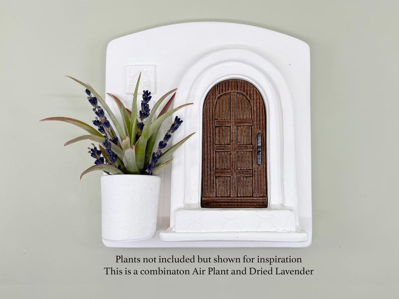 May include: A white wall decor piece featuring a miniature arched doorway with a brown wooden door. A white planter with a green plant and purple flowers sits to the left. The text at the bottom says, "Plants not included but shown for inspiration. This is a combinaton Air Plant and Dried Lavender."