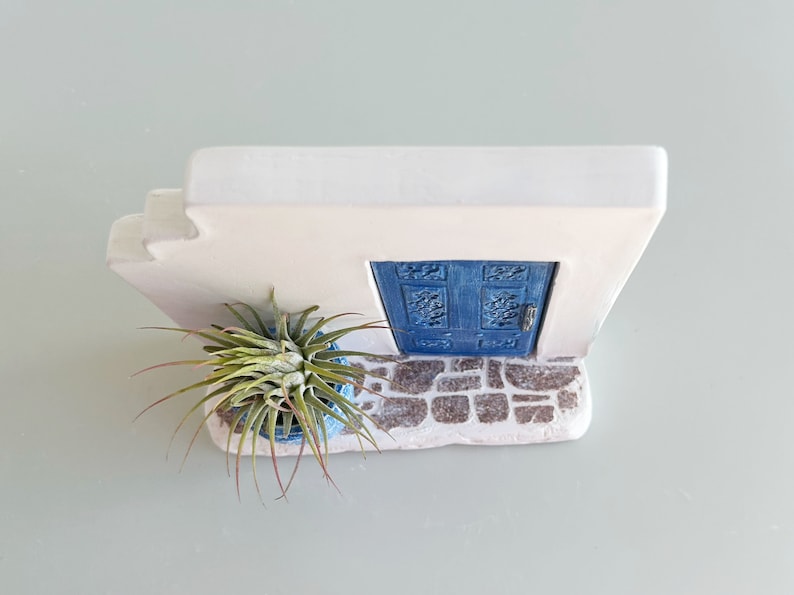 Pode incluir: Uma estrutura de parede branca com uma porta azul de entalhes detalhados e uma base com textura de pedra. Uma planta a&eacute;rea verde e pontiaguda est&aacute; num pequeno vaso azul sobre o ch&atilde;o de pedra em frente &agrave; porta, criando uma cena em miniatura.