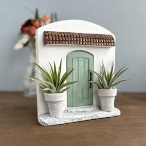 Pu&ograve; includere: Un edificio in miniatura in stucco bianco con una porta verde, un tetto in tegole marroni e due piante in vaso. La porta ha una maniglia nera. Le piante hanno lunghe foglie verdi e sono in vasi bianchi.