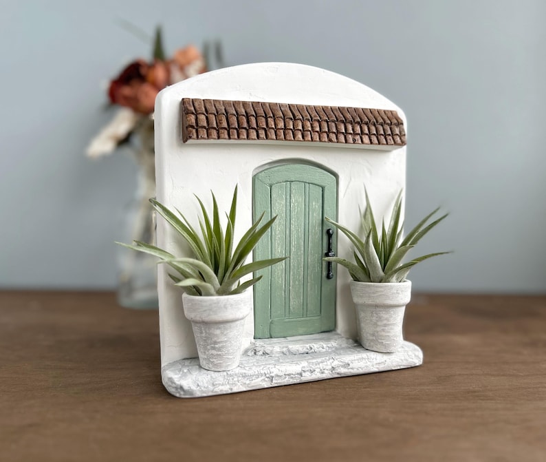 Pu&ograve; includere: Un edificio in miniatura in stucco bianco con una porta verde, un tetto in tegole marroni e due piante in vaso. La porta ha una maniglia nera. Le piante hanno lunghe foglie verdi e sono in vasi bianchi.