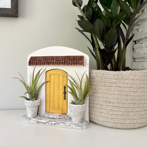 Pu&ograve; includere: Una porta delle fate decorativa con una porta gialla, un tetto marrone e una facciata bianca. Due piccole piante in vaso fiancheggiano la porta. A destra c'&egrave; una grande pianta in vaso in un cesto intrecciato.