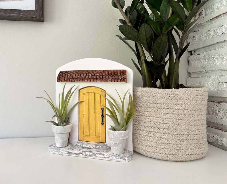Pu&ograve; includere: Una porta delle fate decorativa con una porta gialla, un tetto marrone e una facciata bianca. Due piccole piante in vaso fiancheggiano la porta. A destra c'&egrave; una grande pianta in vaso in un cesto intrecciato.