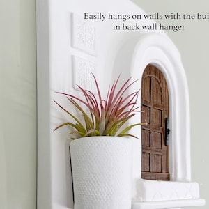 May include: White wall-mounted planter with a textured pot holding a plant with red and green leaves. A miniature wooden door is set into the wall. Text: Easily hangs on walls with the built in back wall hanger.