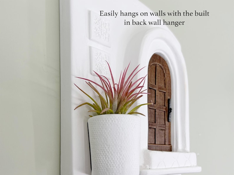 May include: White wall-mounted planter with a textured pot holding a plant with red and green leaves. A miniature wooden door is set into the wall. Text: Easily hangs on walls with the built in back wall hanger.