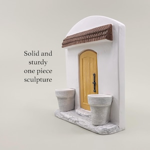 Pu&ograve; includere: Una scultura bianca in miniatura di una facciata di edificio con una porta gialla, un tetto marrone e due fioriere grigie. Il testo "Solid and sturdy one piece sculpture" &egrave; visibile.