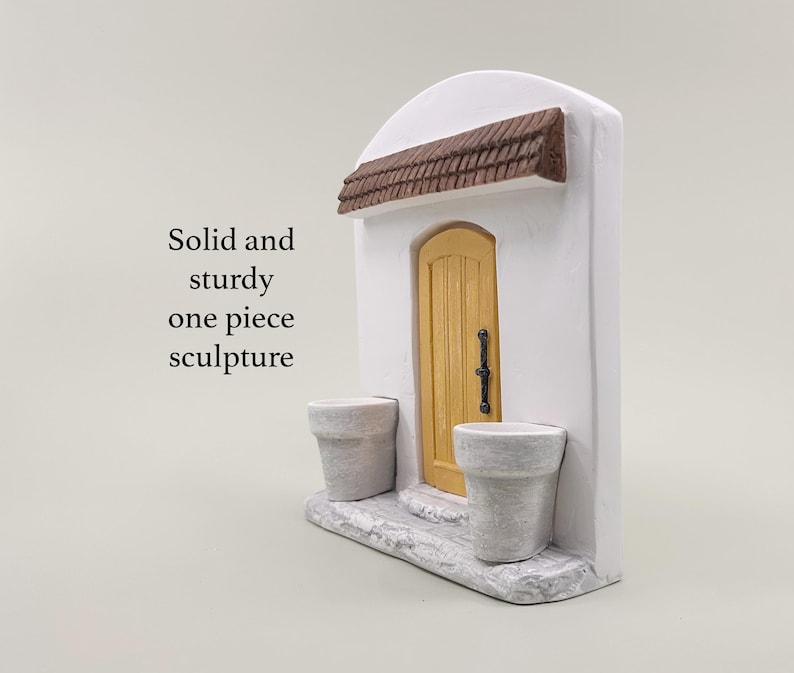 Pu&ograve; includere: Una scultura bianca in miniatura di una facciata di edificio con una porta gialla, un tetto marrone e due fioriere grigie. Il testo "Solid and sturdy one piece sculpture" &egrave; visibile.