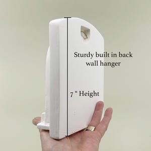 May include: A white, arched wall hanging with a sturdy built-in back wall hanger. The height of the wall hanging is 7 inches. The wall hanging is held by a hand.