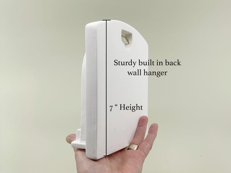 May include: A white, arched wall hanging with a sturdy built-in back wall hanger. The height of the wall hanging is 7 inches. The wall hanging is held by a hand.