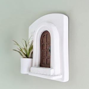 May include: A white wall-mounted decorative piece featuring a miniature arched doorway with a brown wooden door. A small white planter with a green plant is attached to the left side of the doorway. The piece is designed to resemble a Mediterranean style.