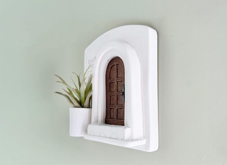 May include: A white wall-mounted decorative piece featuring a miniature arched doorway with a brown wooden door. A small white planter with a green plant is attached to the left side of the doorway. The piece is designed to resemble a Mediterranean style.