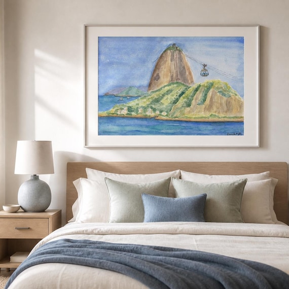 A framed watercolor painting depicting Sugarloaf Mountain in Rio de Janeiro, Brazil. The artwork features the iconic granite peak, lush green hills, and a cable car suspended by wires against a blue sky, set above a calm blue ocean.