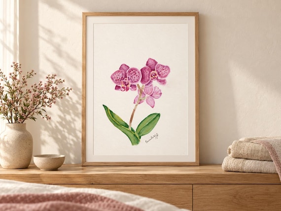 A framed watercolor painting of a magenta orchid with spotted petals and green leaves. The artwork features a signature in the bottom right corner and is displayed in a light wood frame on a wooden surface.