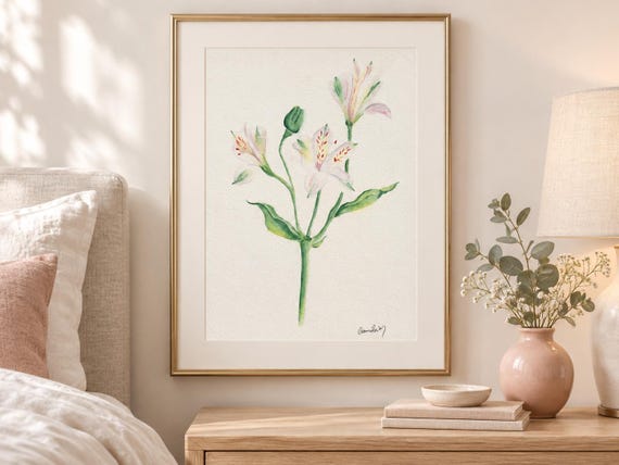 A framed watercolor painting featuring a single stem of white and pale pink alstroemeria flowers with green leaves and a small bud. The artwork is enclosed in a thin gold frame and includes a signature in the bottom right corner.