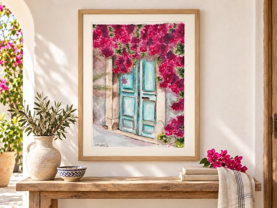 A framed watercolor painting depicting a pair of light blue wooden doors surrounded by vibrant magenta bougainvillea flowers. The artwork features a signature in the bottom left corner and is displayed in a light wood frame.